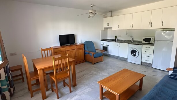 A modern living room with a dining area, a kitchen with white cabinets, a ceiling fan, and a blue sofa.