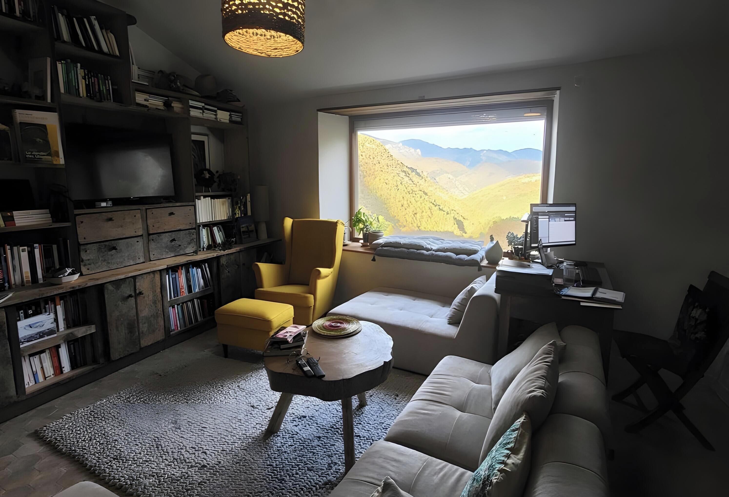 Un salon chaleureux avec une bibliothèque, un fauteuil jaune, un canapé et une grande fenêtre donnant sur les montagnes.