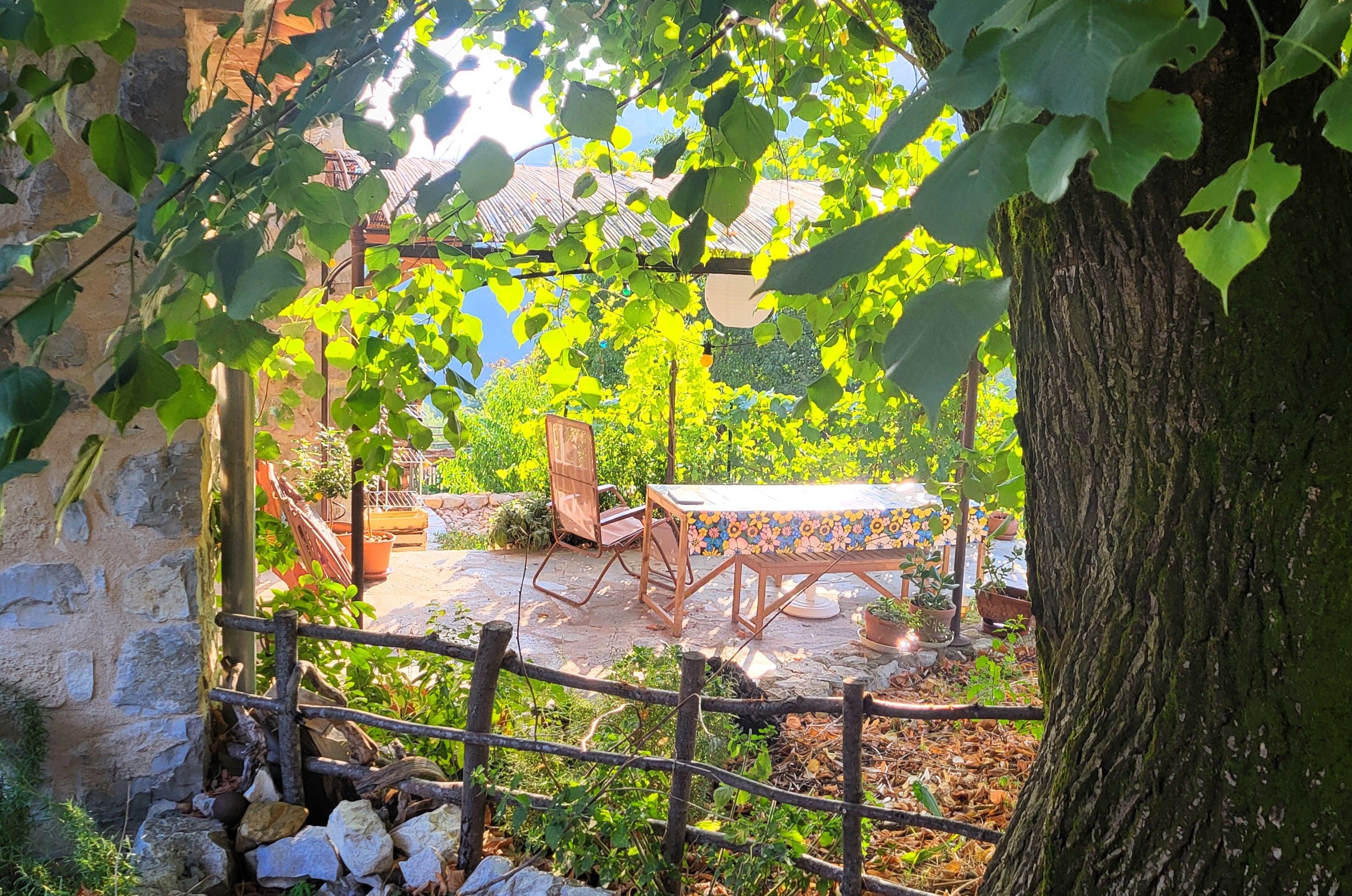 A rustic outdoor seating area with a wooden table and chairs, surrounded by greenery and a stone wall.