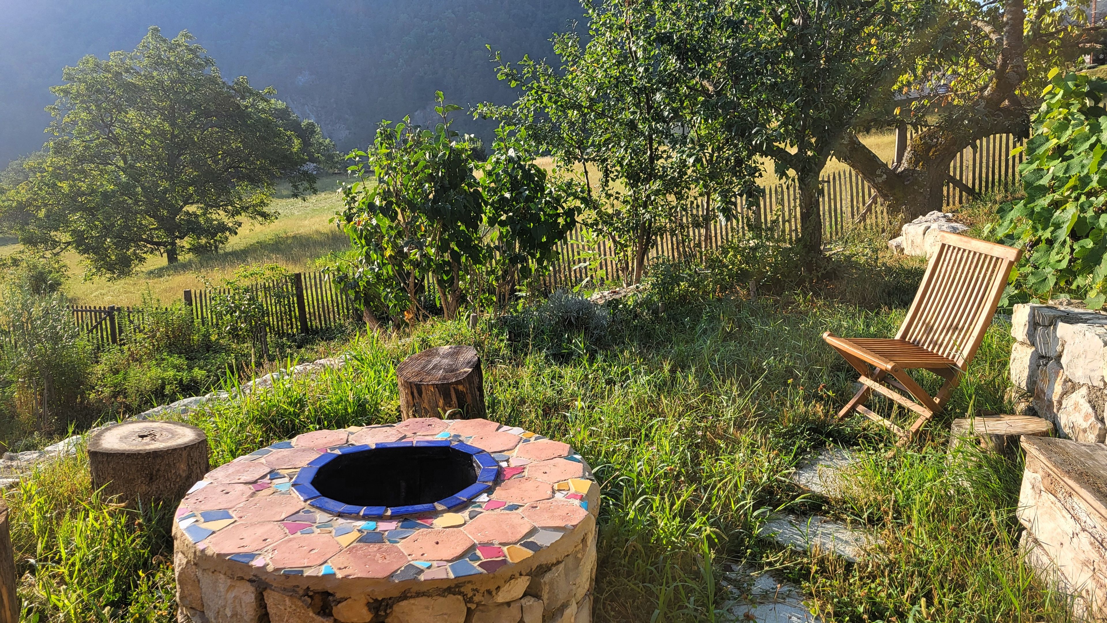 Um jardim com área de estar ao ar livre coberta por mosaicos, um banco de madeira e uma lareira rústica.