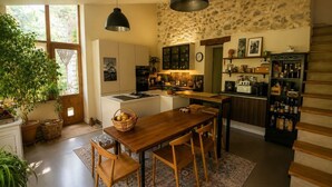 A modern kitchen with a central island, wooden dining table, and chairs, surrounded by potted plants and a stone wall.