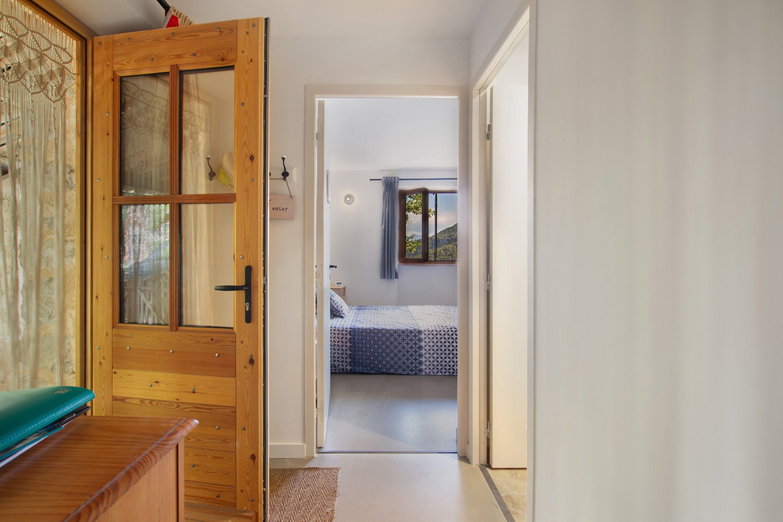 A hallway leading to a bedroom with a bed, a wooden door, and a view of the outdoors.