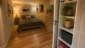 A cozy bedroom with a wooden bed, a nightstand with a lamp, a bookshelf with books and decorative items, and a view of the outdoors through a window.