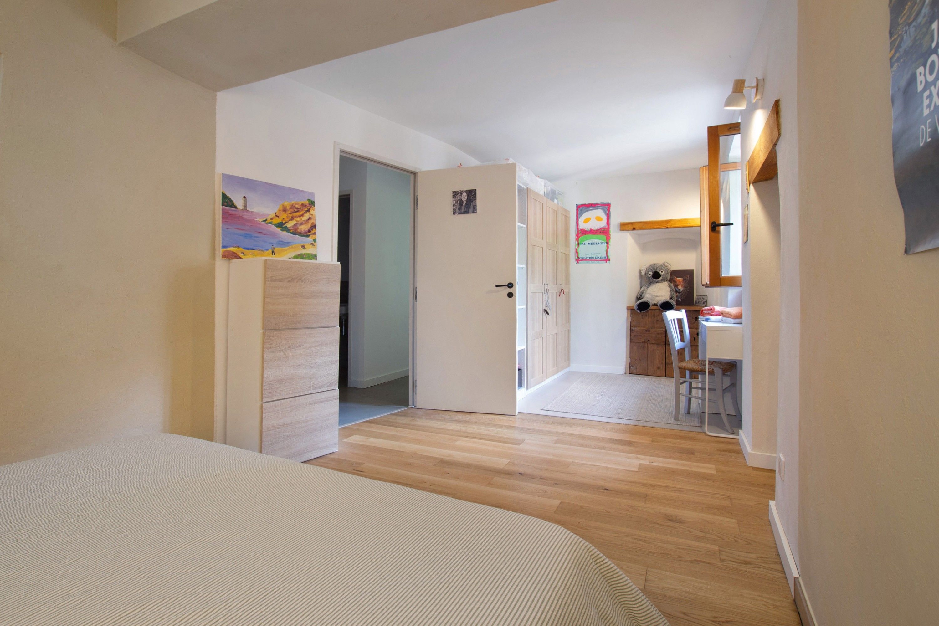 A room with a bed, a door leading to a kitchen, and a painting on the wall.