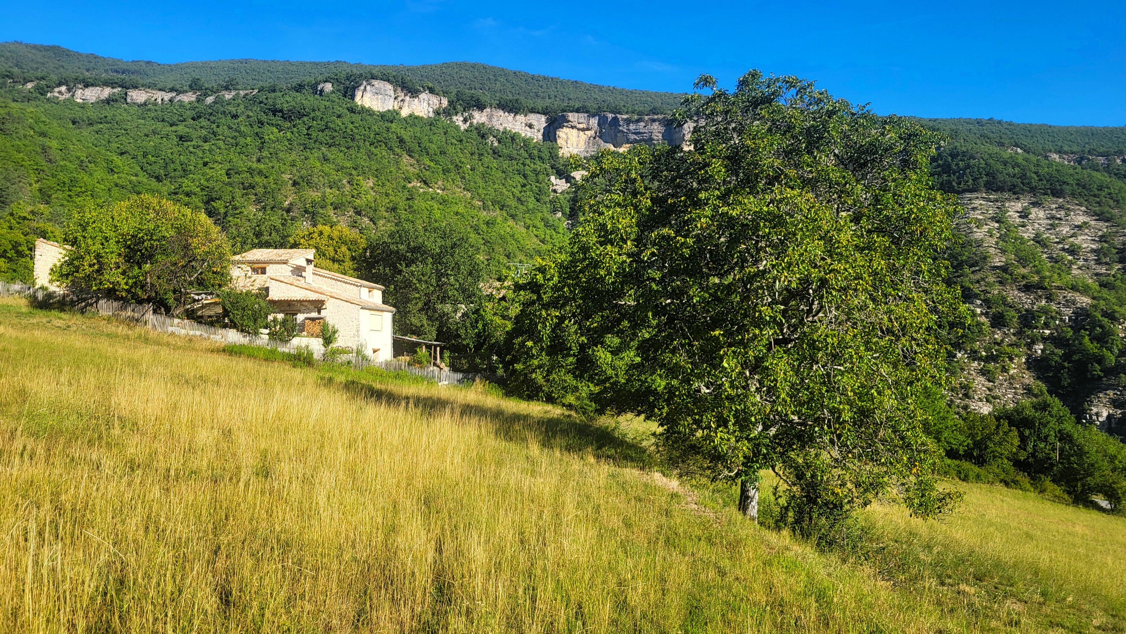 Una casa rural con fachada de piedra, rodeada por un campo cubierto de hierba y una ladera boscosa.