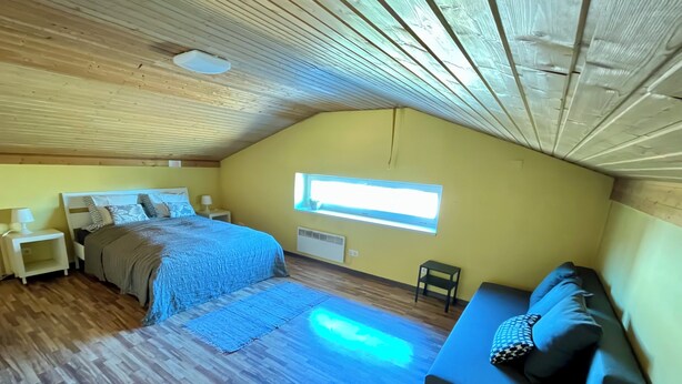 A bedroom with a bed, bedside tables, a window, a sofa, and a wooden ceiling.