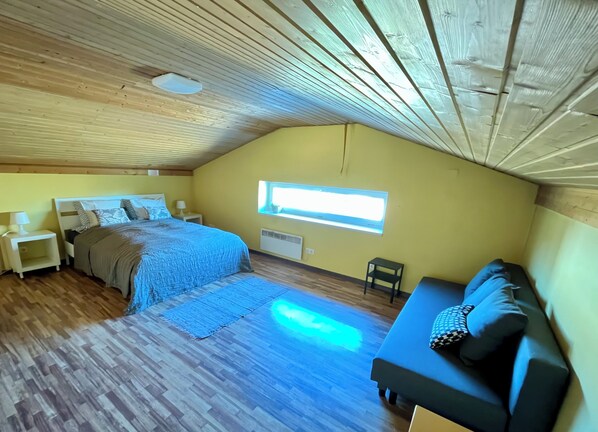A bedroom with a bed, bedside tables, a window, a sofa, and a wooden ceiling.
