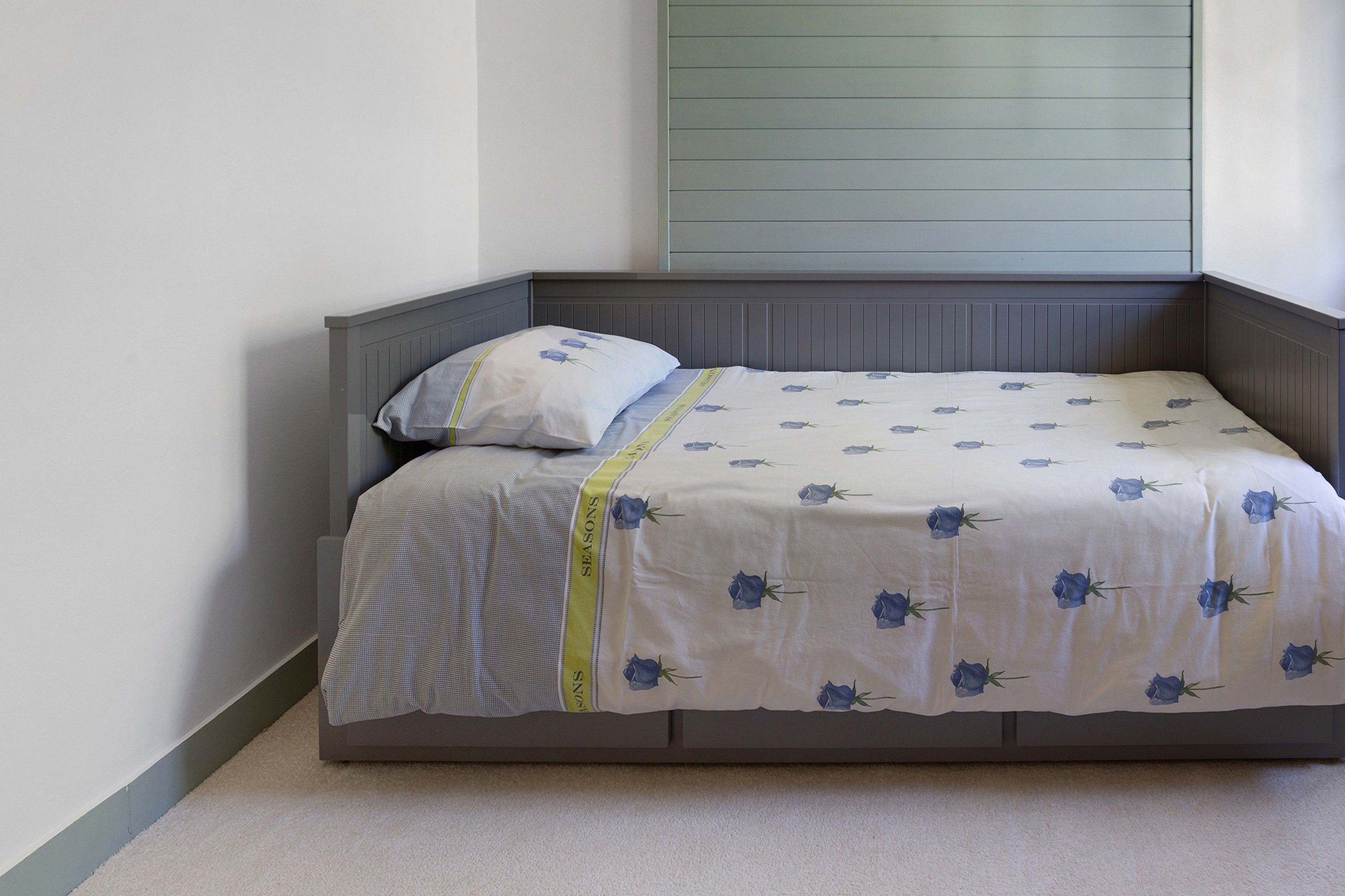 Un lit simple avec une tête de lit et un pied de lit gris, une couvre-lit blanche ornée de motifs floraux bleus et un oreiller au motif similaire.