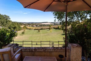 Pátio com mesa e cadeiras, com vista para uma paisagem rural com campos e uma aldeia.