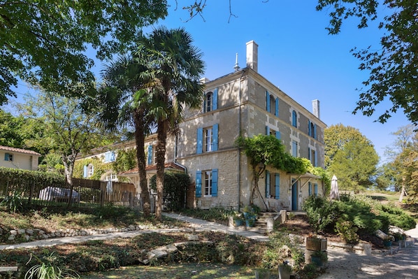 A two-story stone building with blue shutters, surrounded by lush greenery and a stone pathway.