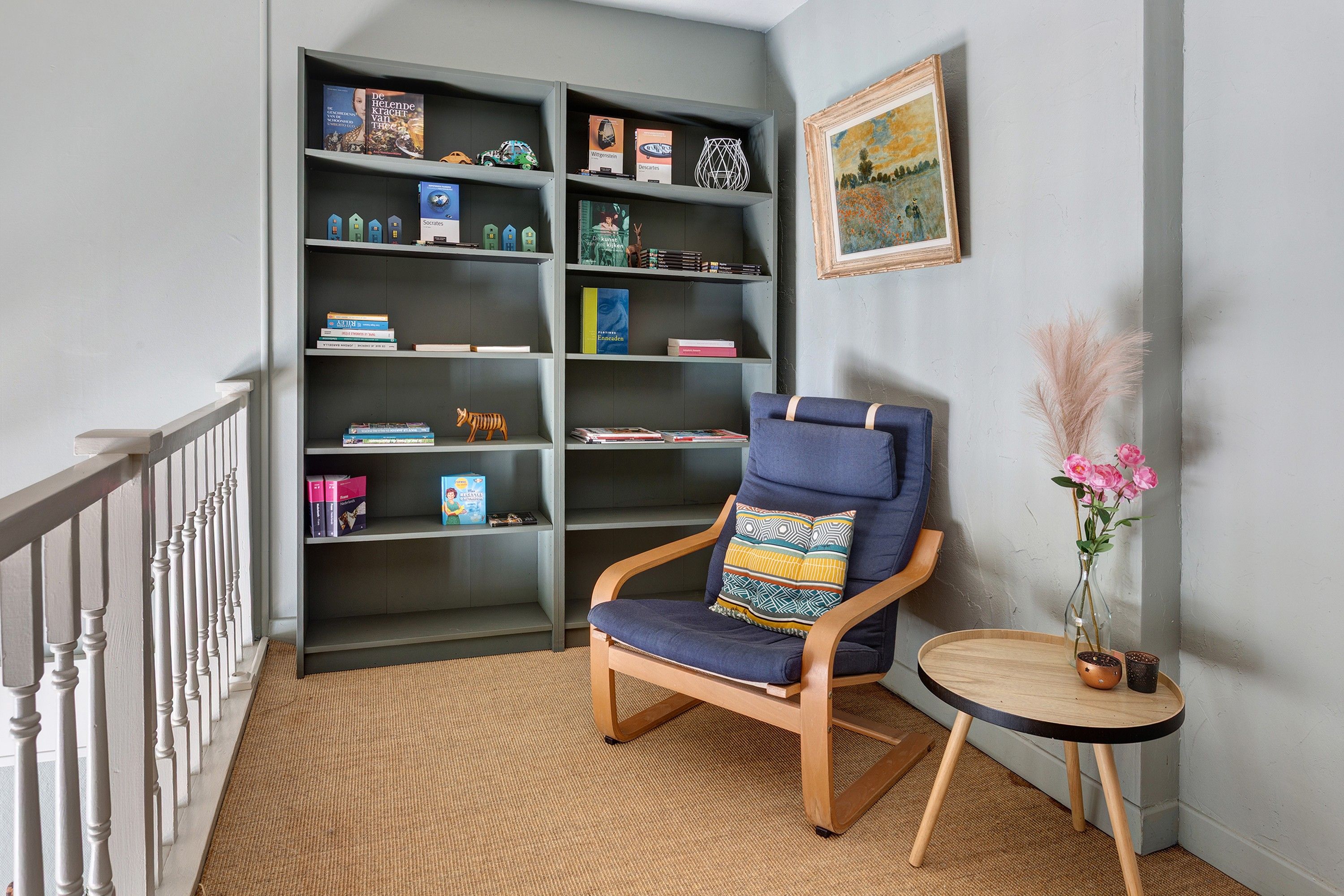 A corner room with a bookshelf, a chair, a small table with a vase, and a painting on the wall.