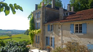 A two-story building with a red-tiled roof, blue shutters, and a garden area with a table and chairs.