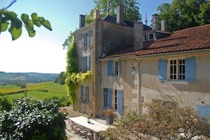 A two-story building with a red-tiled roof and blue shutters, surrounded by greenery and a view of the countryside.