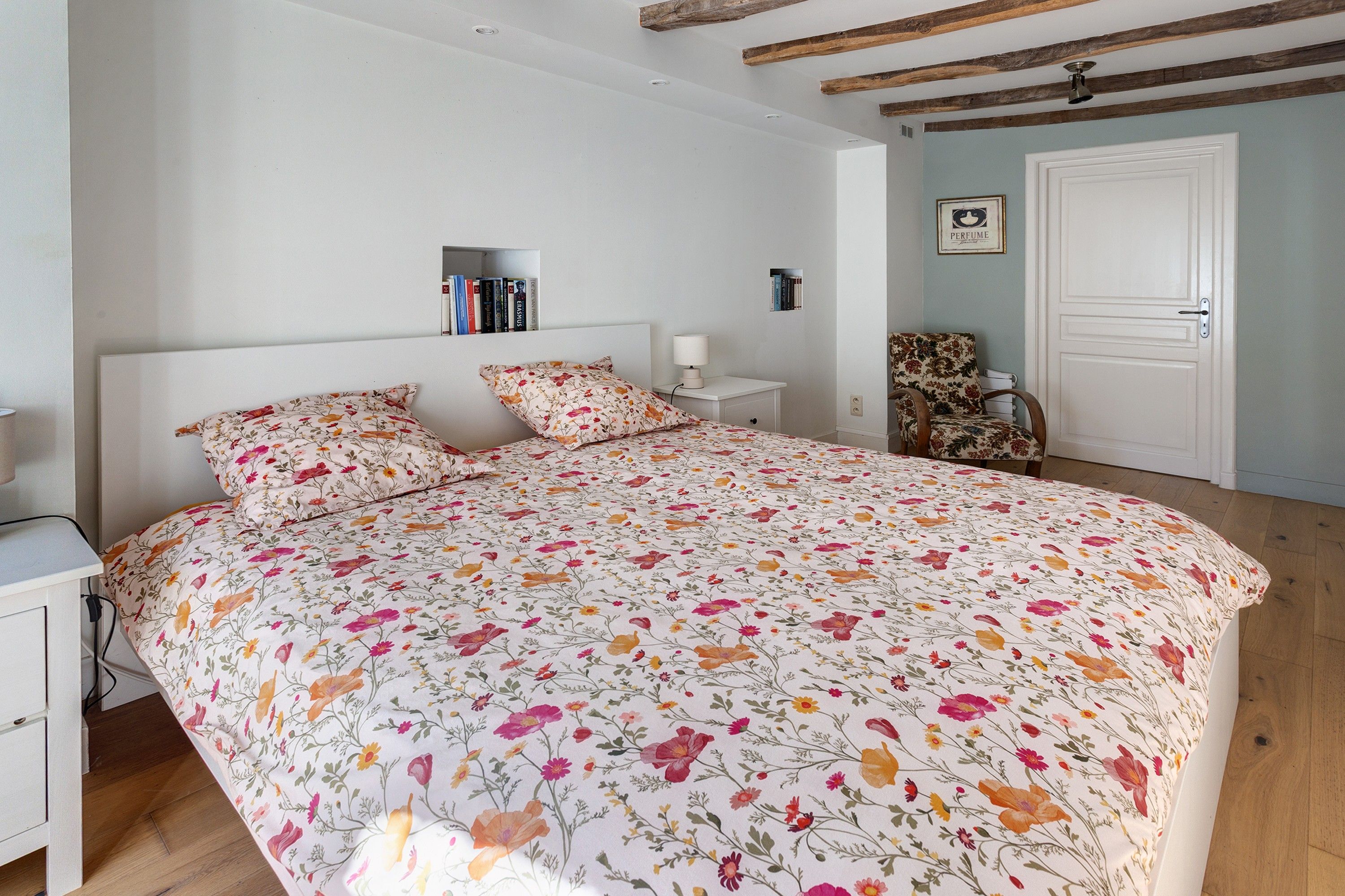 A bedroom with a large bed, a bedside table, a chair, a bookshelf, and a door.