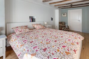 Quarto com uma cama grande, mesa de cabeceira, uma cadeira, estante de livros e uma porta.