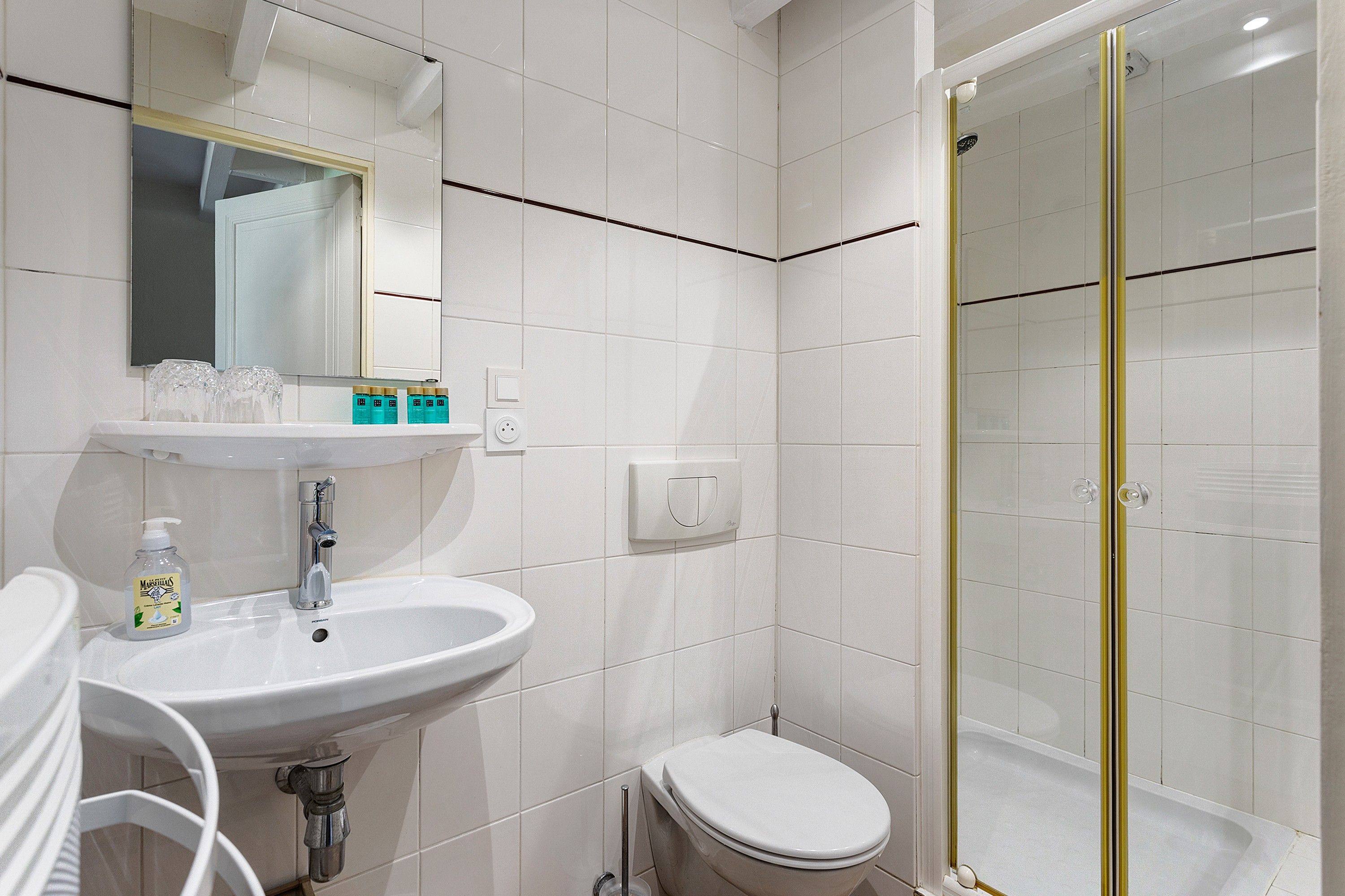 Une salle de bain avec un lavabo blanc, un miroir et des toilettes, une douche en verre et un mur recouvert de carreaux blancs.