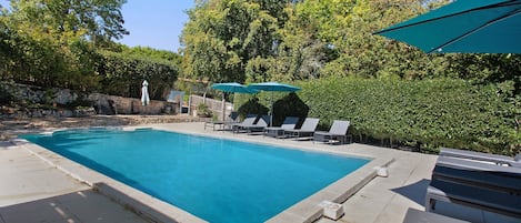 A rectangular swimming pool with a tiled edge, surrounded by lounge chairs and umbrellas, set against a backdrop of trees and a stone wall.