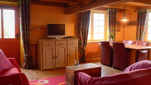 Ein gemütliches Wohnzimmer mit einem Fernseher auf einem Schrank, einem Esstisch mit Stühlen und einem Fenster mit Vorhängen.