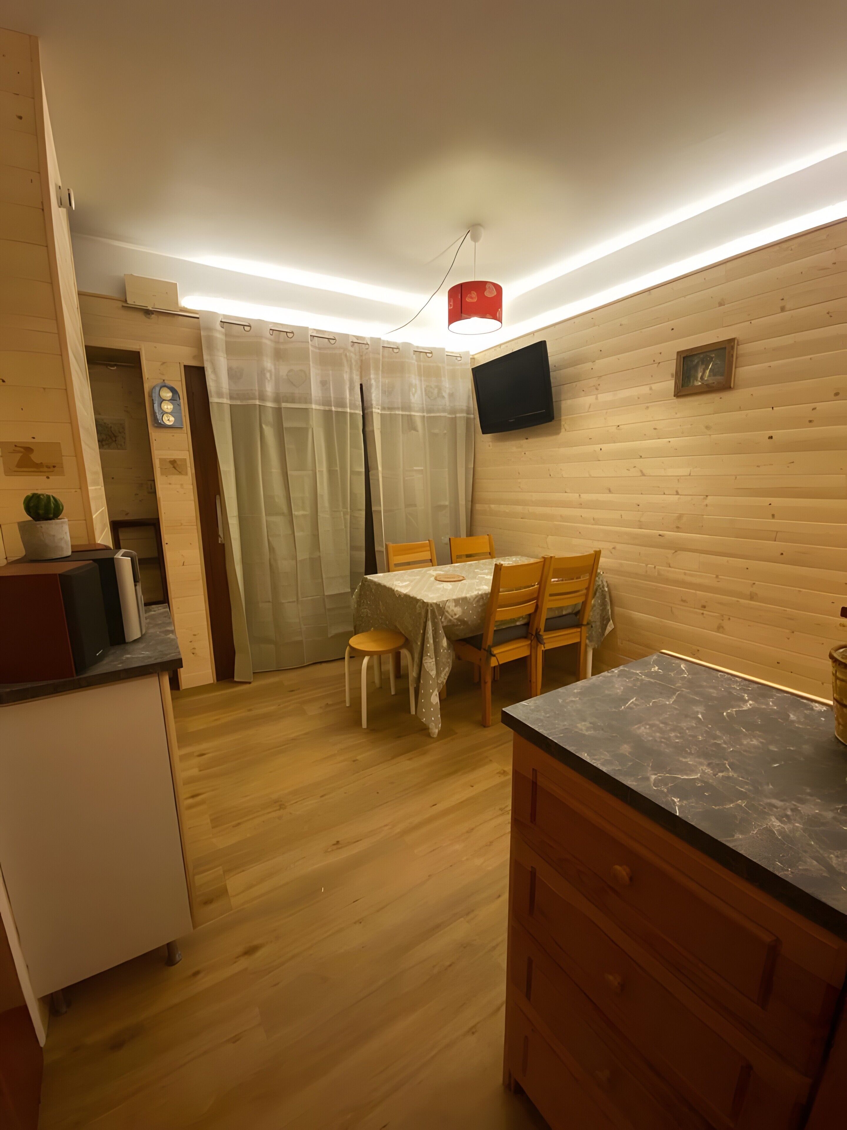 A compact kitchen with wooden cabinets, a dining table with chairs, a small refrigerator, and a flat-screen TV mounted on the wall.