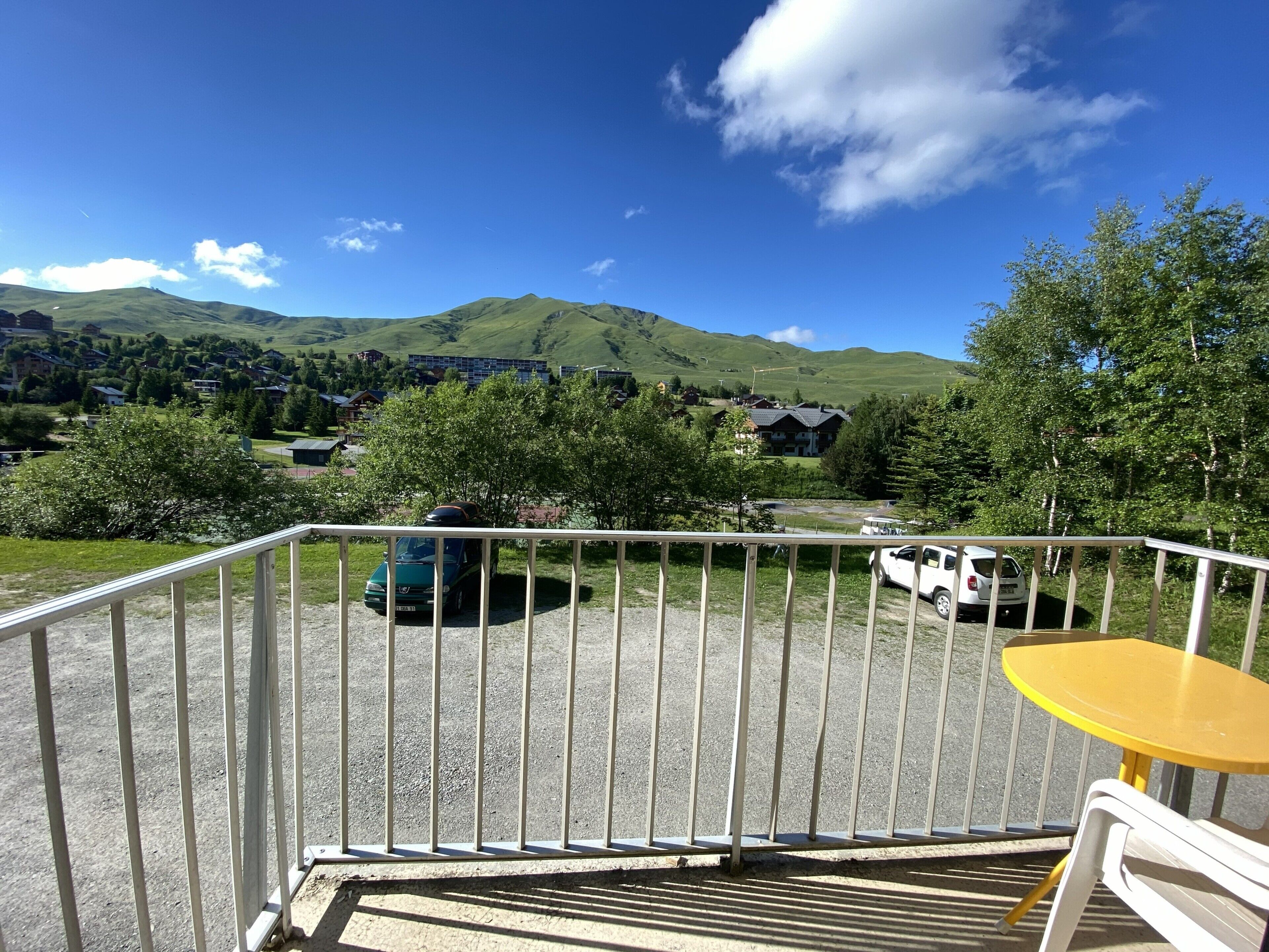 Un balcon doté d’une rambarde en métal, une table jaune et une vue sur un parking où l’on aperçoit des voitures et des maisons au loin.
