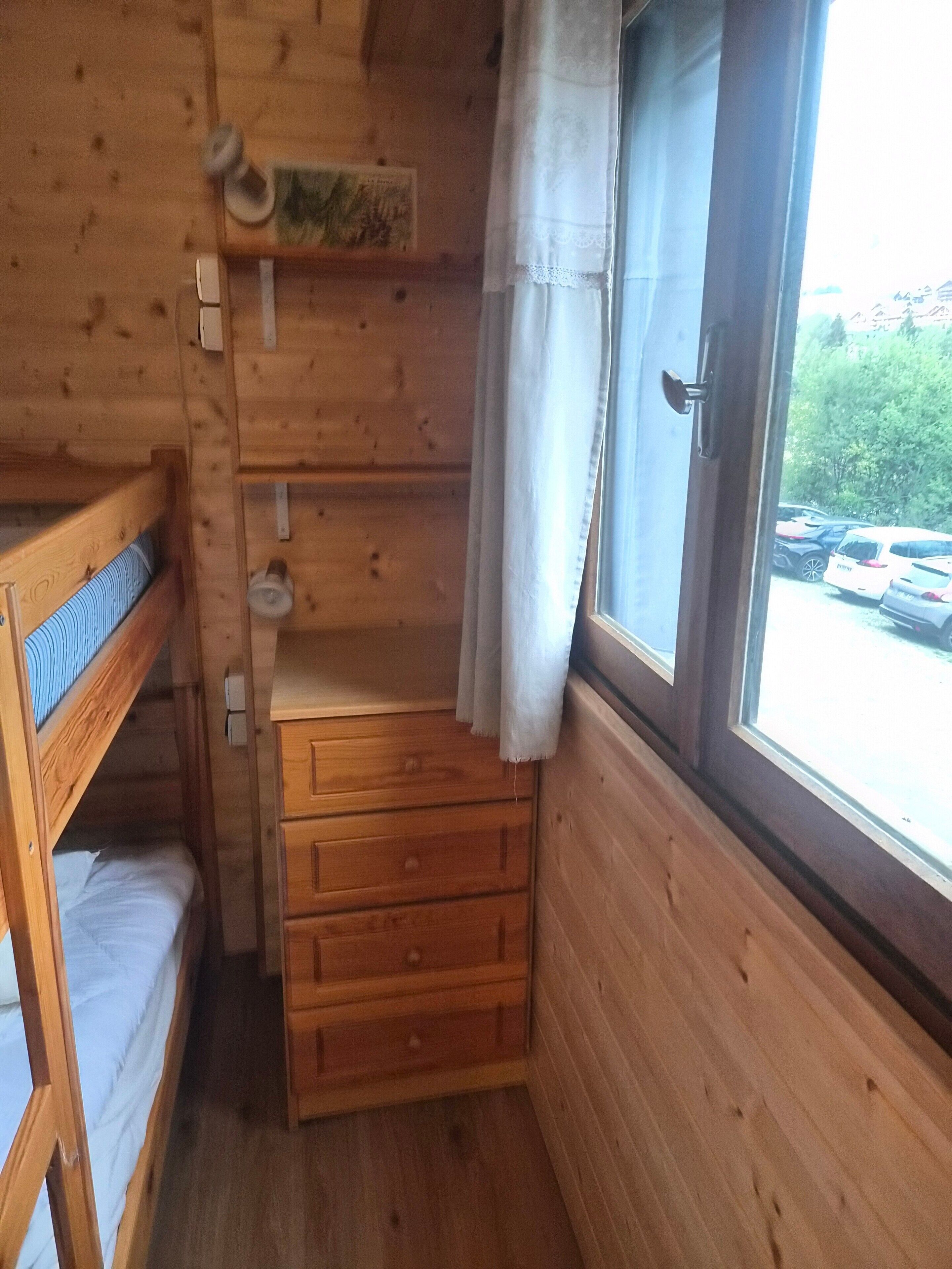 A wooden cabin room with bunk beds, a small wooden dresser, and a window with curtains.