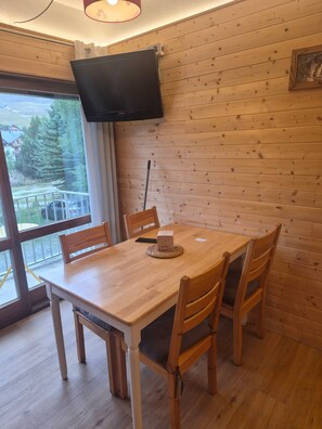 Ein Zimmer mit Holzwänden, ausgestattet mit einem Tisch, Stühlen, einem an der Wand befestigten Fernseher und einem Fenster mit Blick auf einen Balkon.