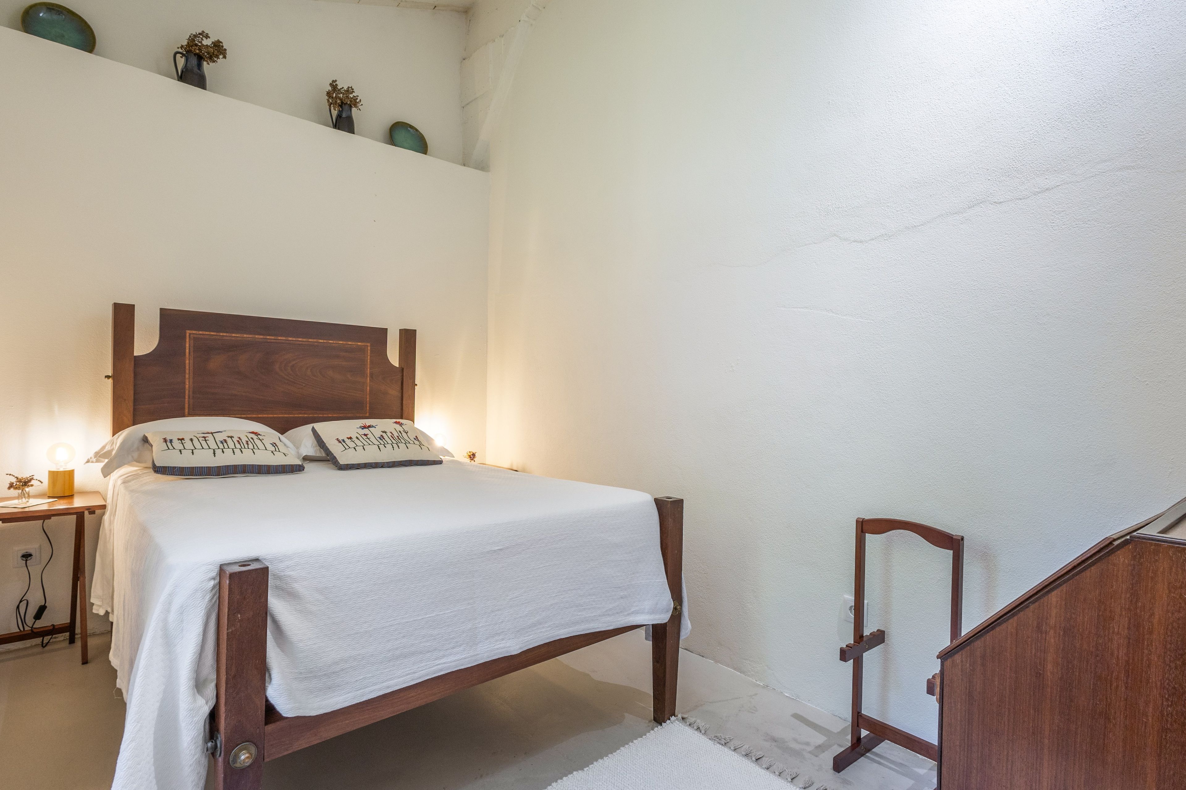A bedroom with a wooden bed, white bedding, a nightstand with a lamp, and a small shelf with decorative items.