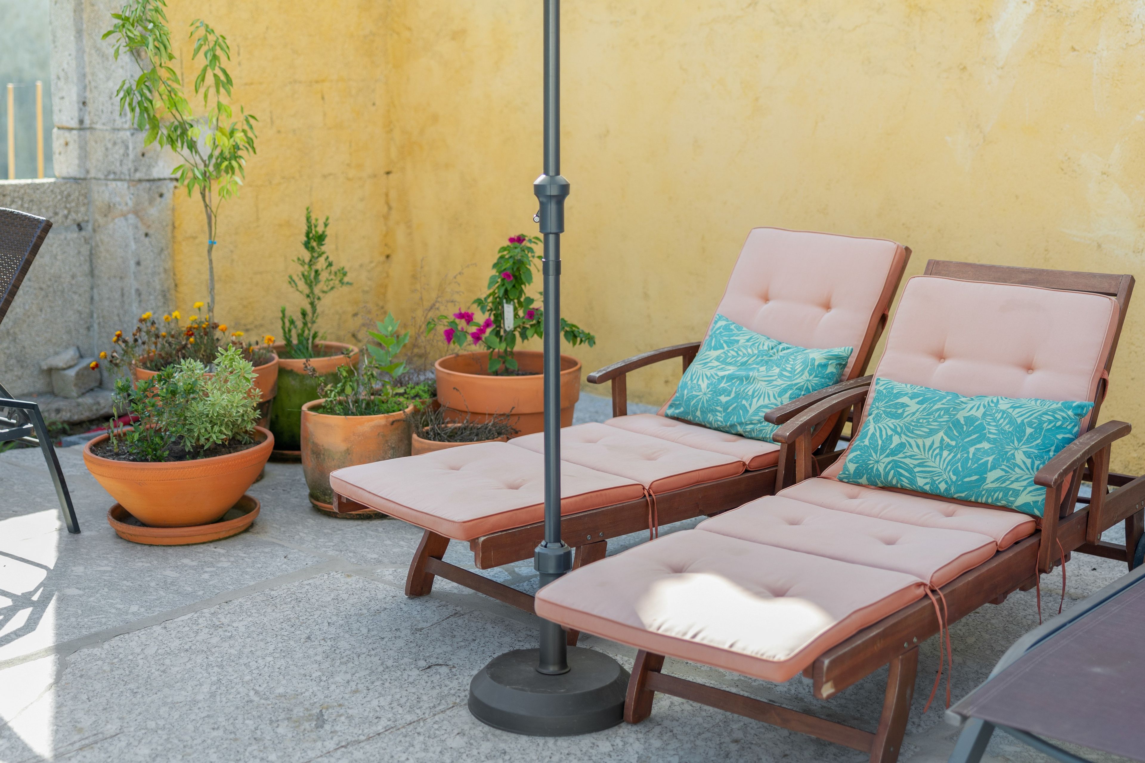 A patio with two cushioned lounge chairs, a metal umbrella, and various potted plants.