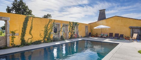 En swimmingpool med en gul vägg och stenvalv, omgiven av solstolar och en klar himmel.
