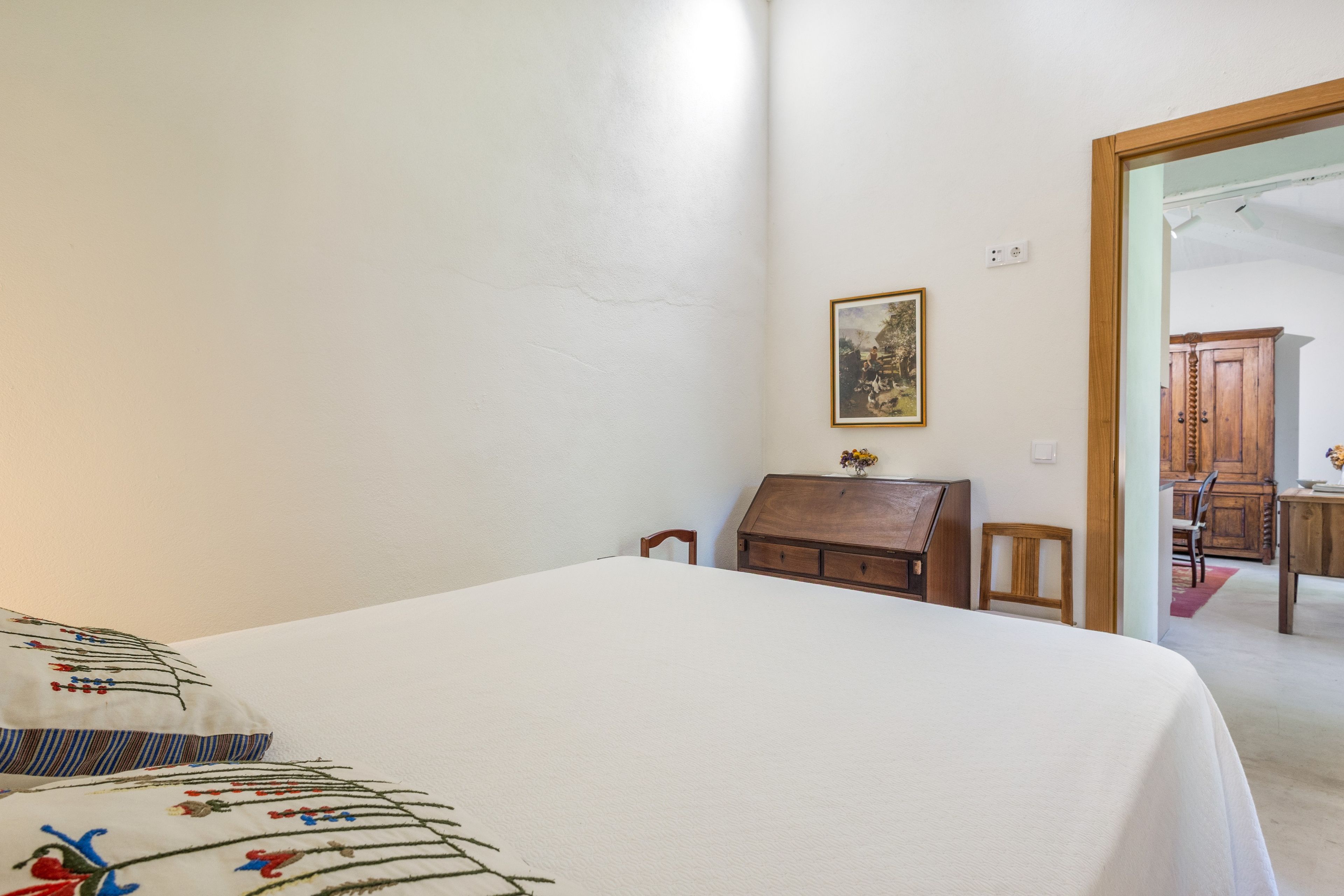 A bedroom with a bed, a wooden chest of drawers, a small table with a vase, and a framed picture on the wall.