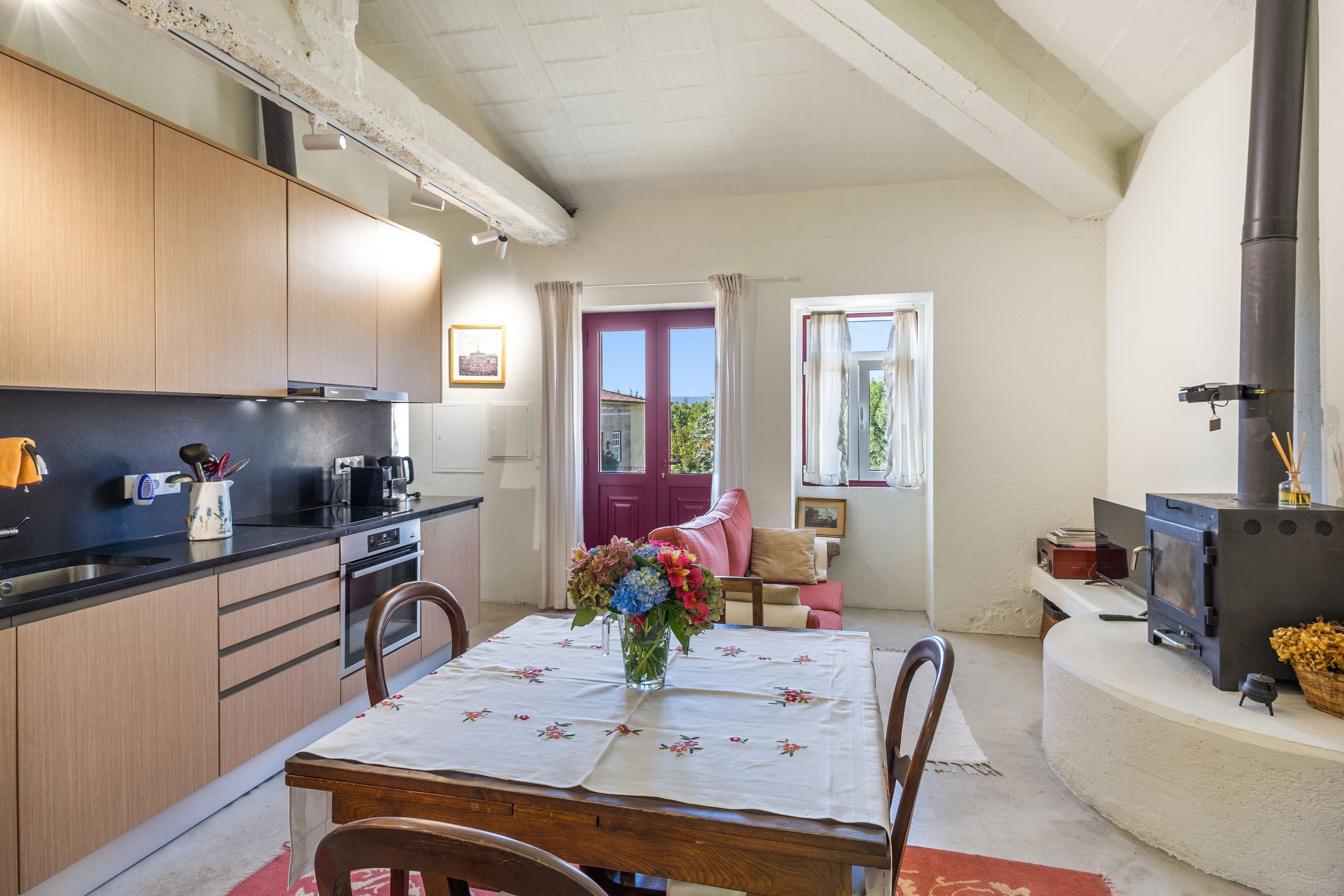 A cozy kitchen with wooden cabinets, a dining table with a floral tablecloth, and a red door leading to a balcony.