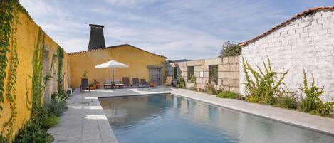 Una piscina con sillas de descanso y una mesa, rodeada por un edificio amarillo y una pared blanca.
