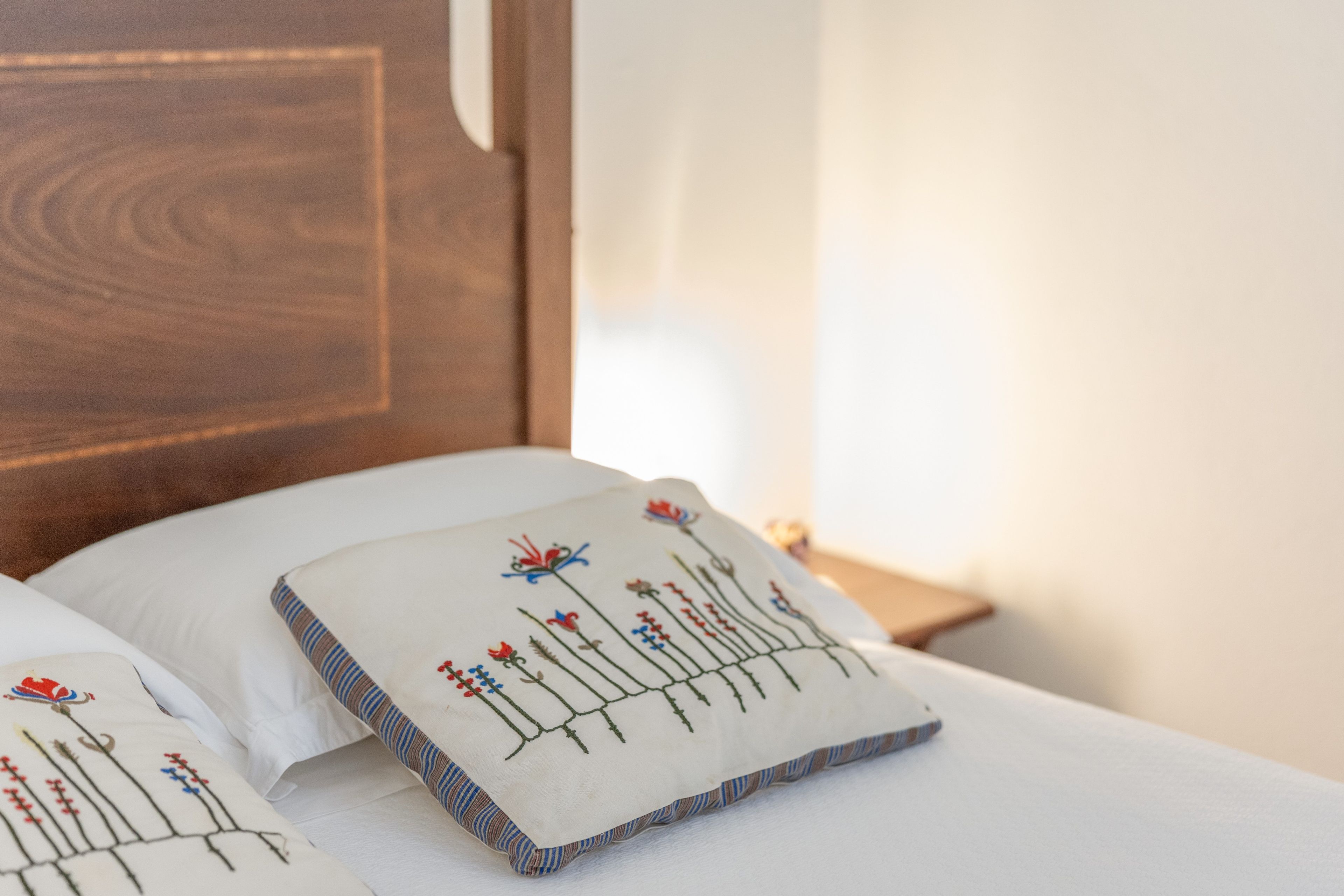Een bed met een wit kussen met geborduurde bloemen en bladeren, een houten hoofdbord en een nachtkastje met een lamp.