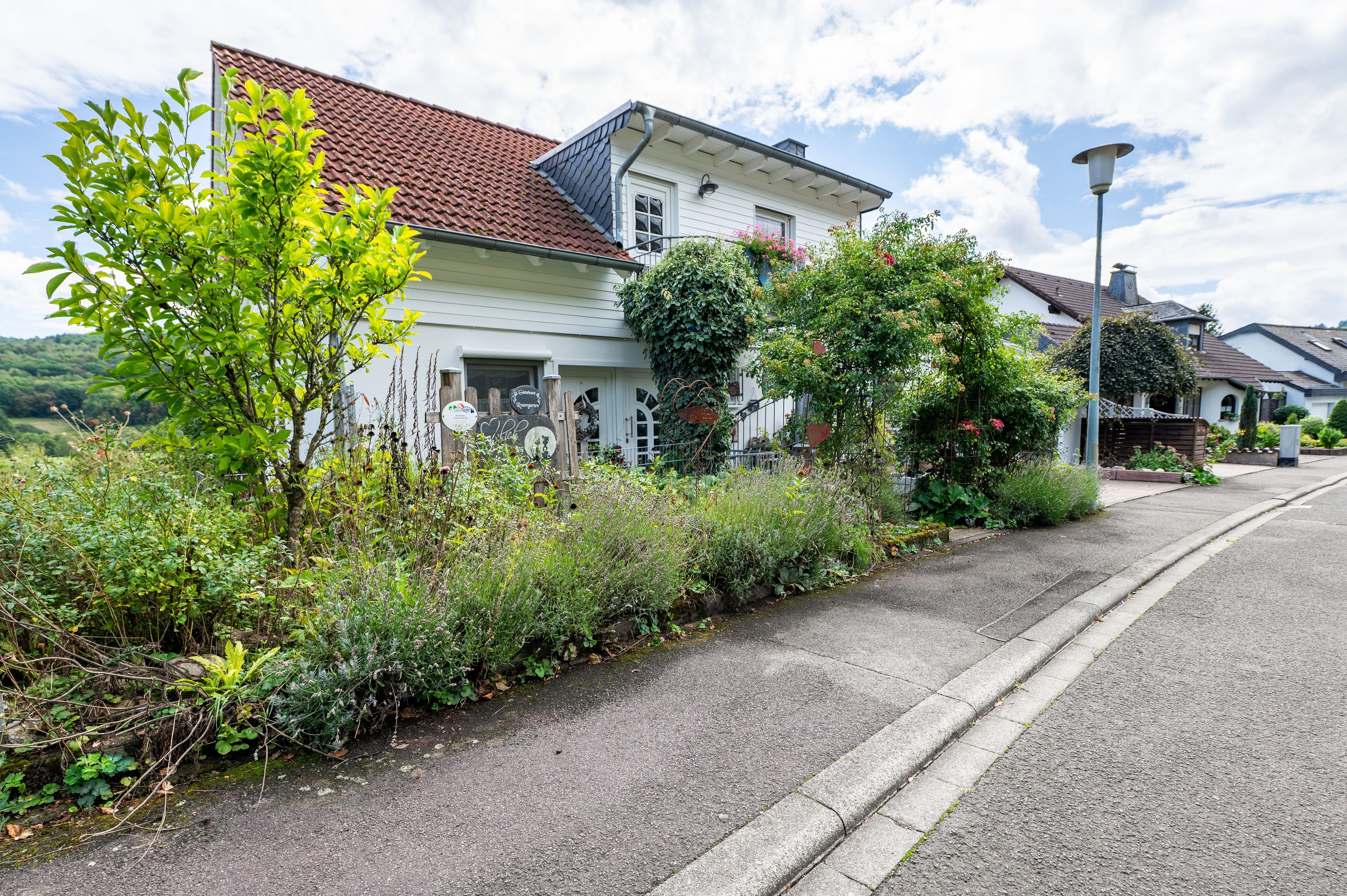 Eine Wohnstraße mit einem weißen Haus, das ein rotes Ziegeldach, einen Garten mit verschiedenen Pflanzen und einen klaren Himmel zeigt.