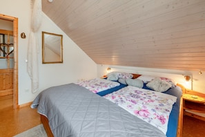 Une chambre avec un plafond en pente, un lit recouvert d’une literie à motifs floraux, une tête de lit en bois et une table de chevet avec une lampe.