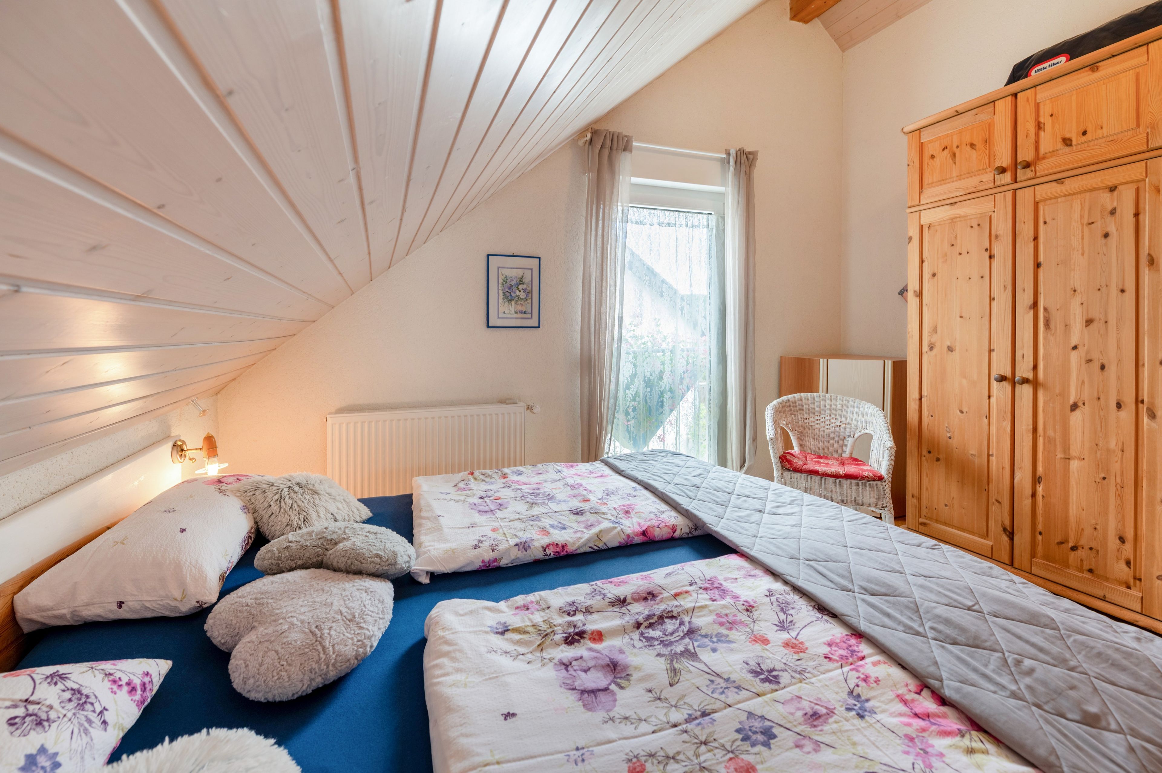A bedroom with a sloped ceiling, a bed with floral bedding, a white chair, and wooden furniture.