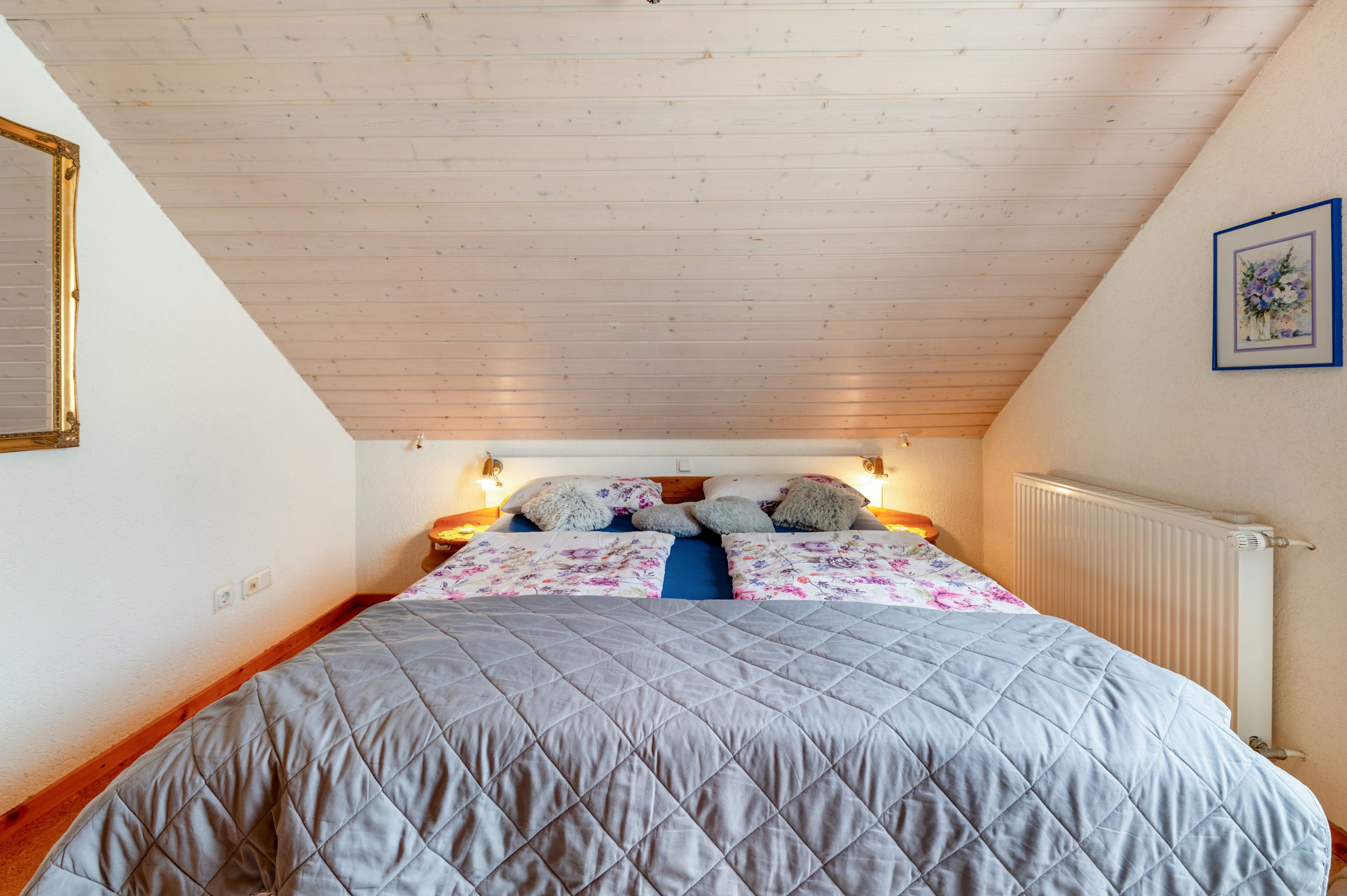 Ein Schlafzimmer mit schrägverdeckter Decke, einem Bett mit gesteppter Auflage, zwei Nachttischlampen und einem gerahmten Bild an der Wand.
