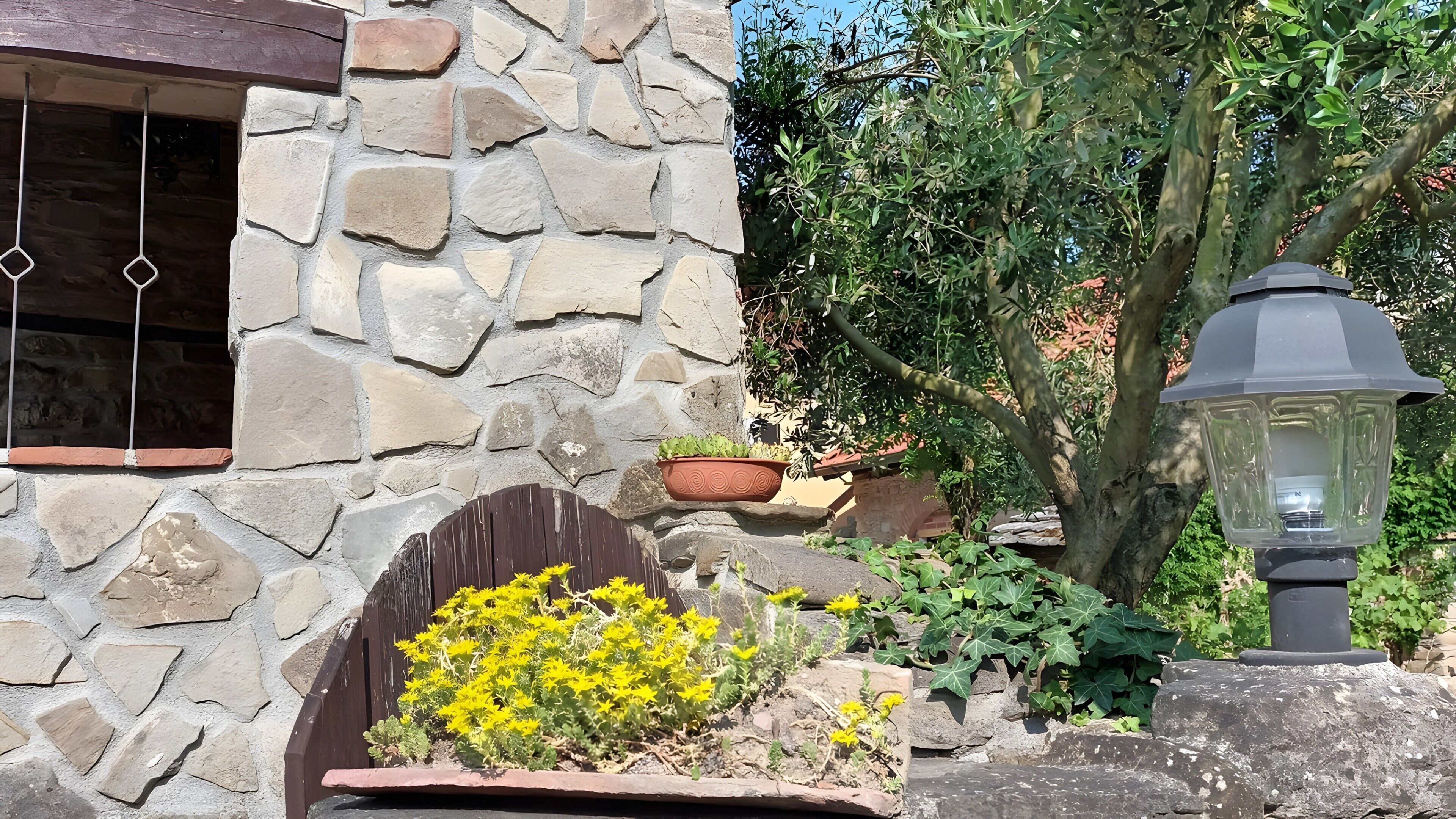 Un muro de piedra con una ventana, un cantero con flores amarillas y una planta en maceta.