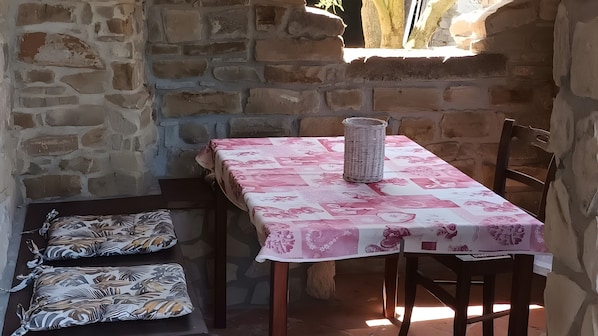A stone-walled room with a table covered in a patterned tablecloth, two cushioned benches, and a woven basket.