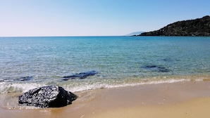 Una playa con agua cristalina, una roca grande en primer plano y un paisaje montañoso al fondo.