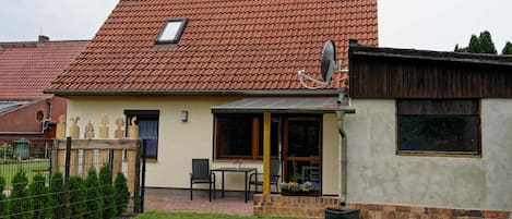 A two-story house with a red-tiled roof, a satellite dish, and a small patio with a table and chairs.