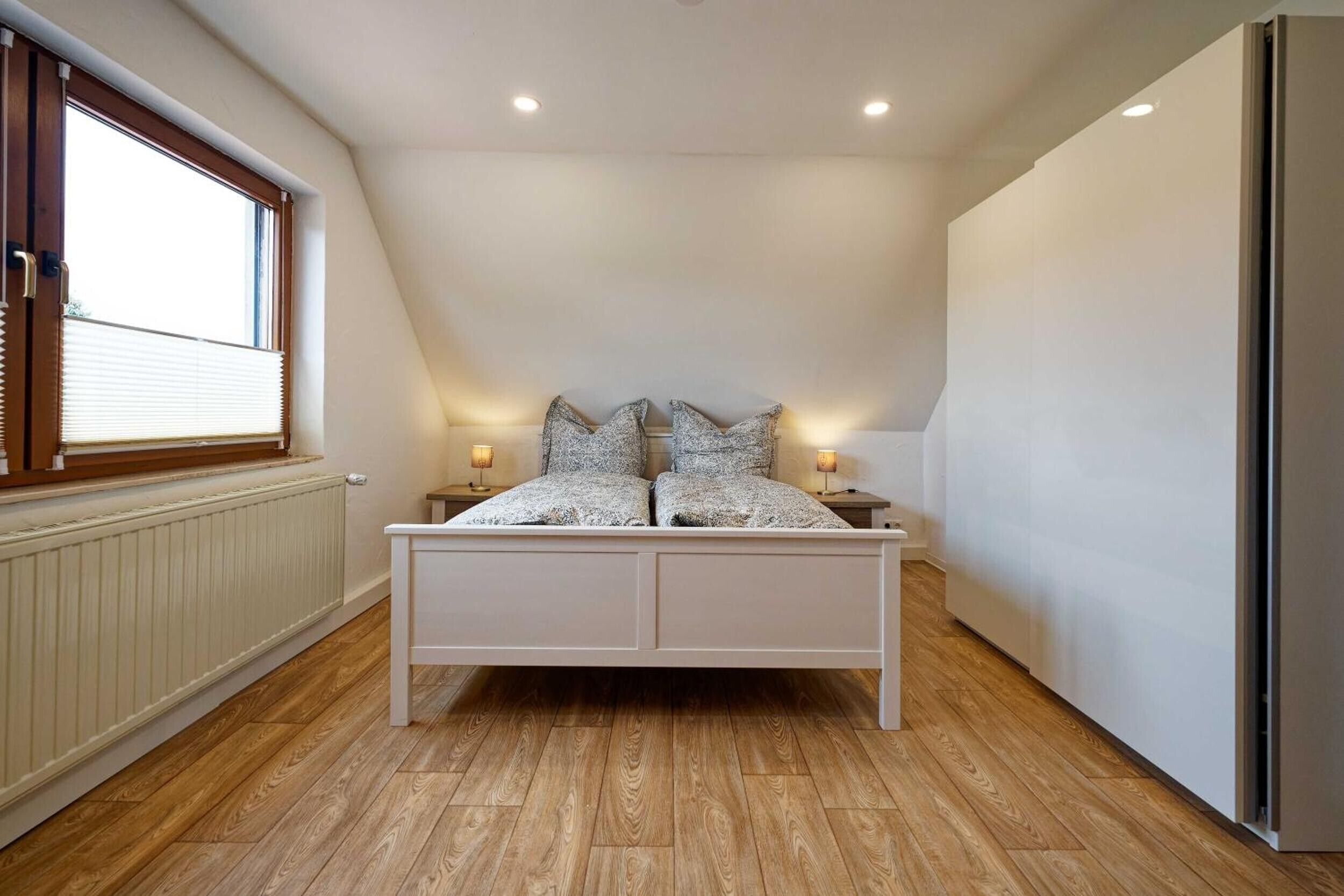 Ein Schlafzimmer mit schrägen Wänden, einem Bett mit gemusterten Textilien, zwei Nachttischlampen und einem Fenster mit Jalousien.