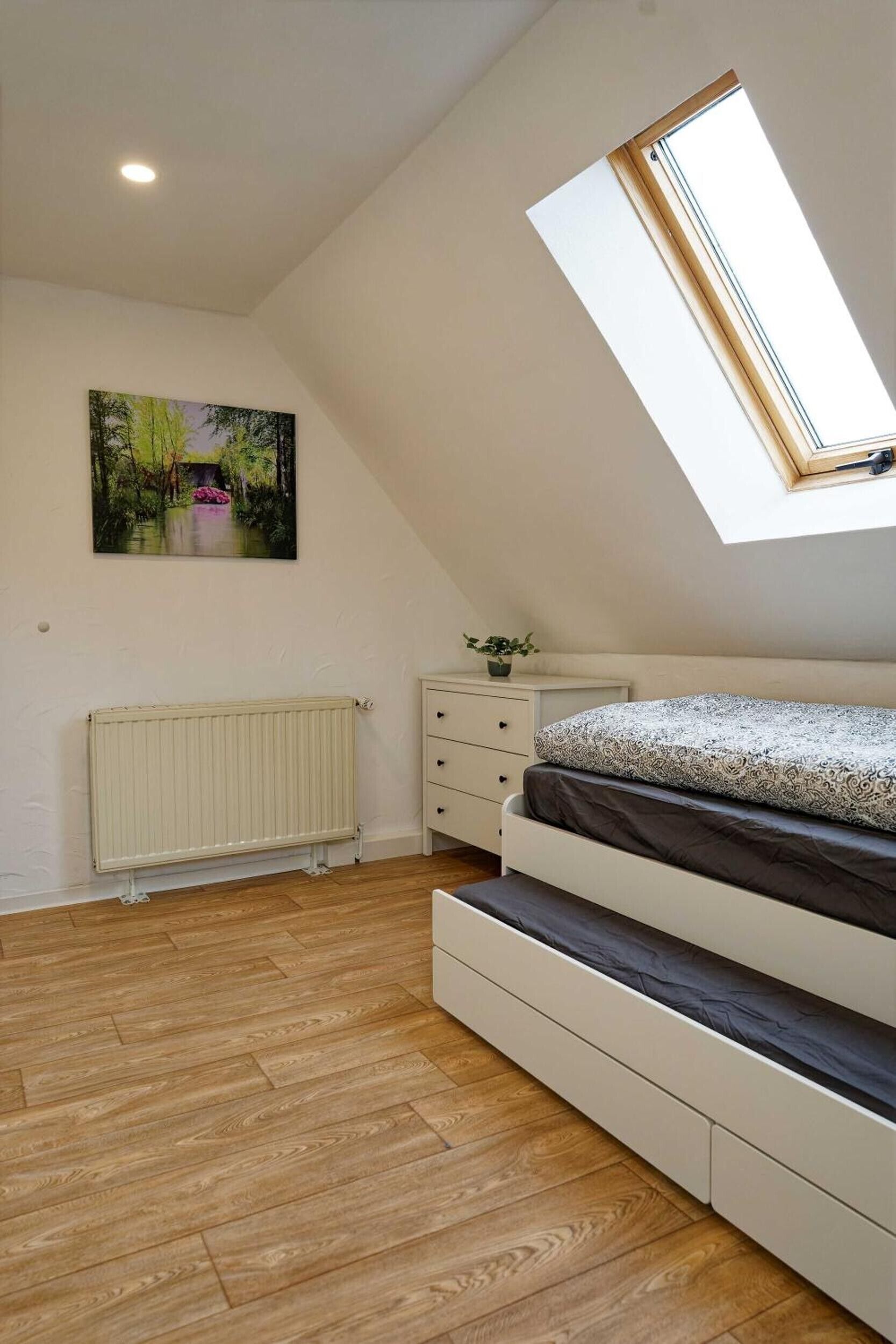 Ein Schlafzimmer mit schrägem Dach, einem Oberlicht, einem Heizkörper, einem Bett mit gemustertem Überwurf, einem Schminktisch und einem Landschaftsbild.