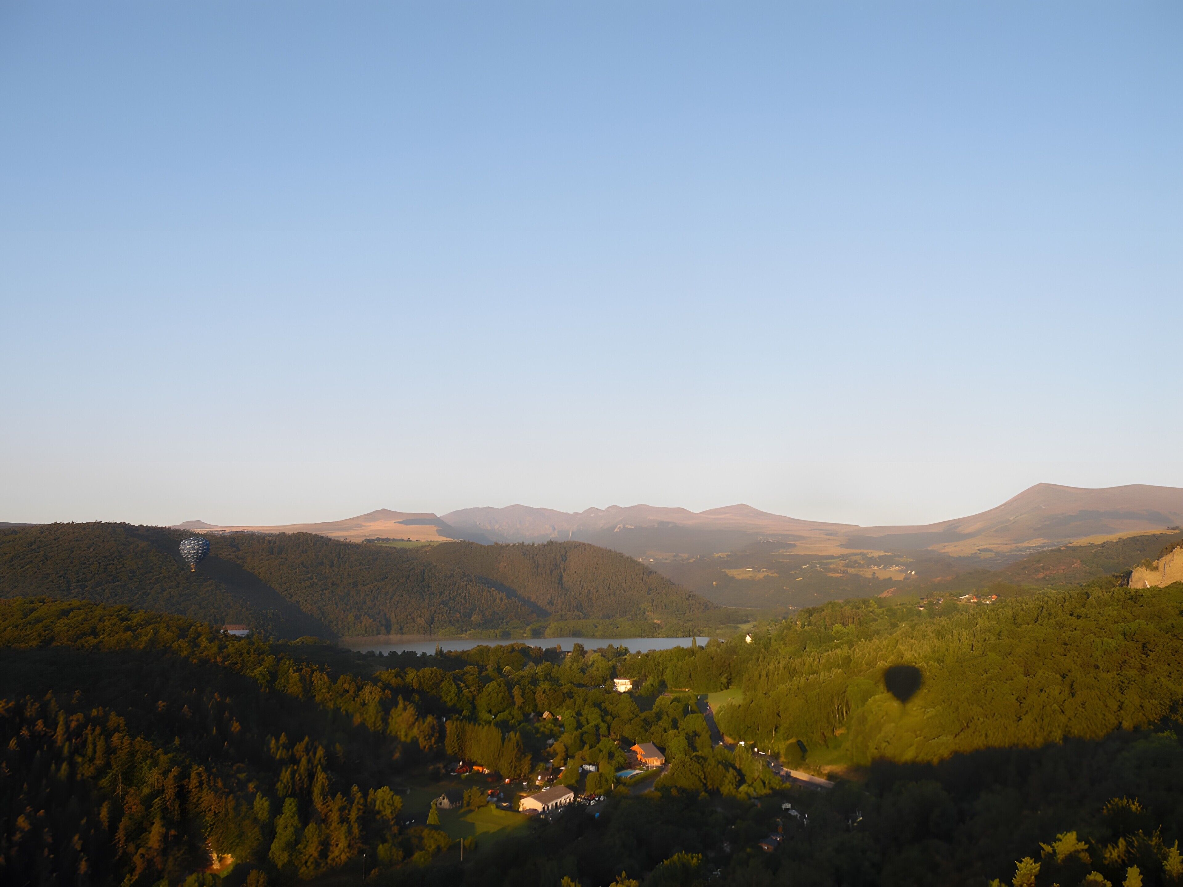 A panoramic view of a valley with a lake, forested hills, and a small settlement.