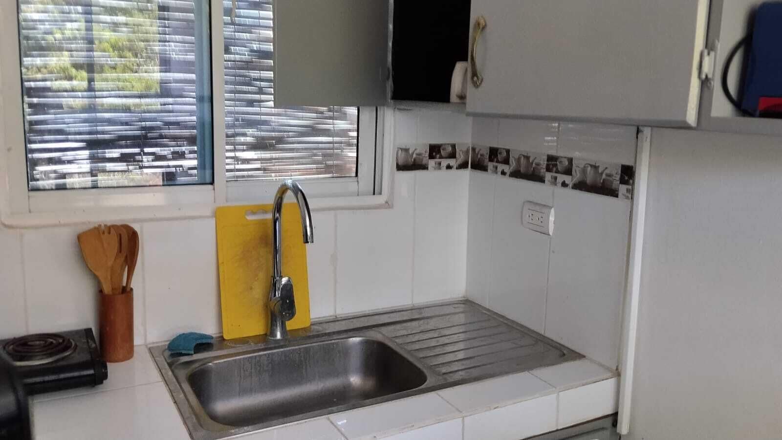 Een keuken met een roestvrijstalen spoelbak, een raam met jaloezieën en een betegelde spatwand.