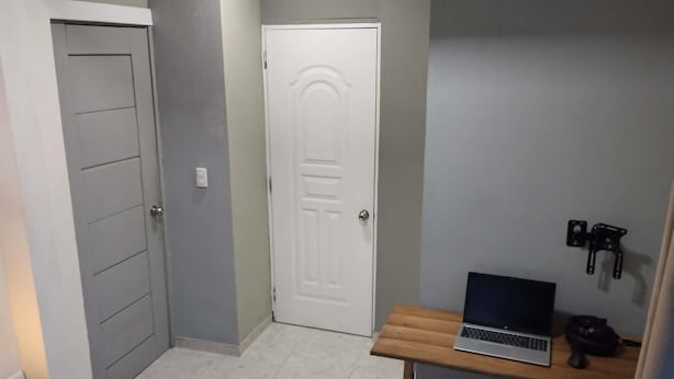 Un pasillo con una puerta, una pequeña mesa con una computadora portátil y una lámpara fijada a la pared.
