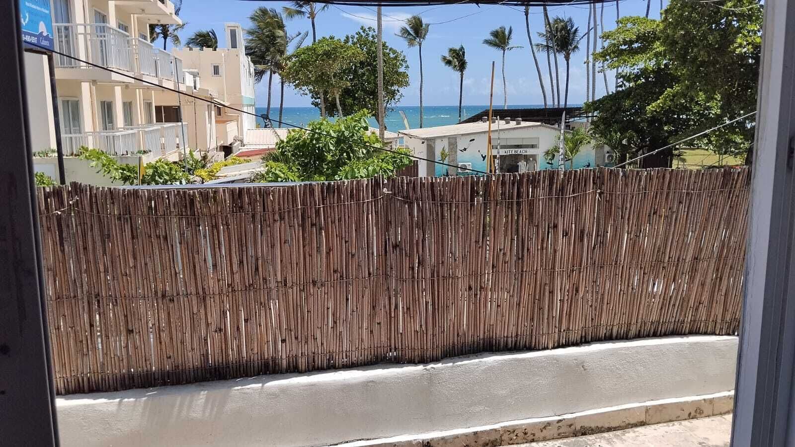 Une vue à travers une fenêtre, avec une clôture en bambou, une plage et des bâtiments en arrière-plan.