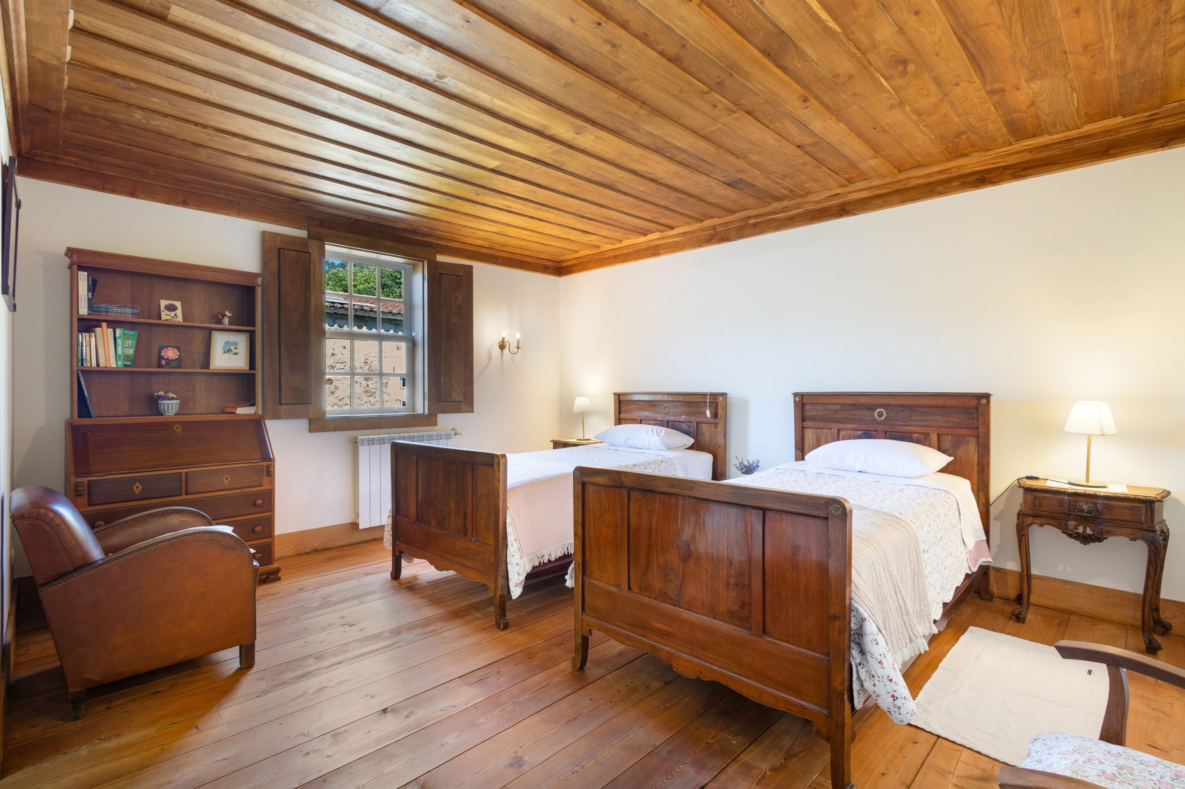 Une pièce avec deux lits en bois, une chaise en bois, une bibliothèque et une fenêtre donnant sur un paysage verdoyant.