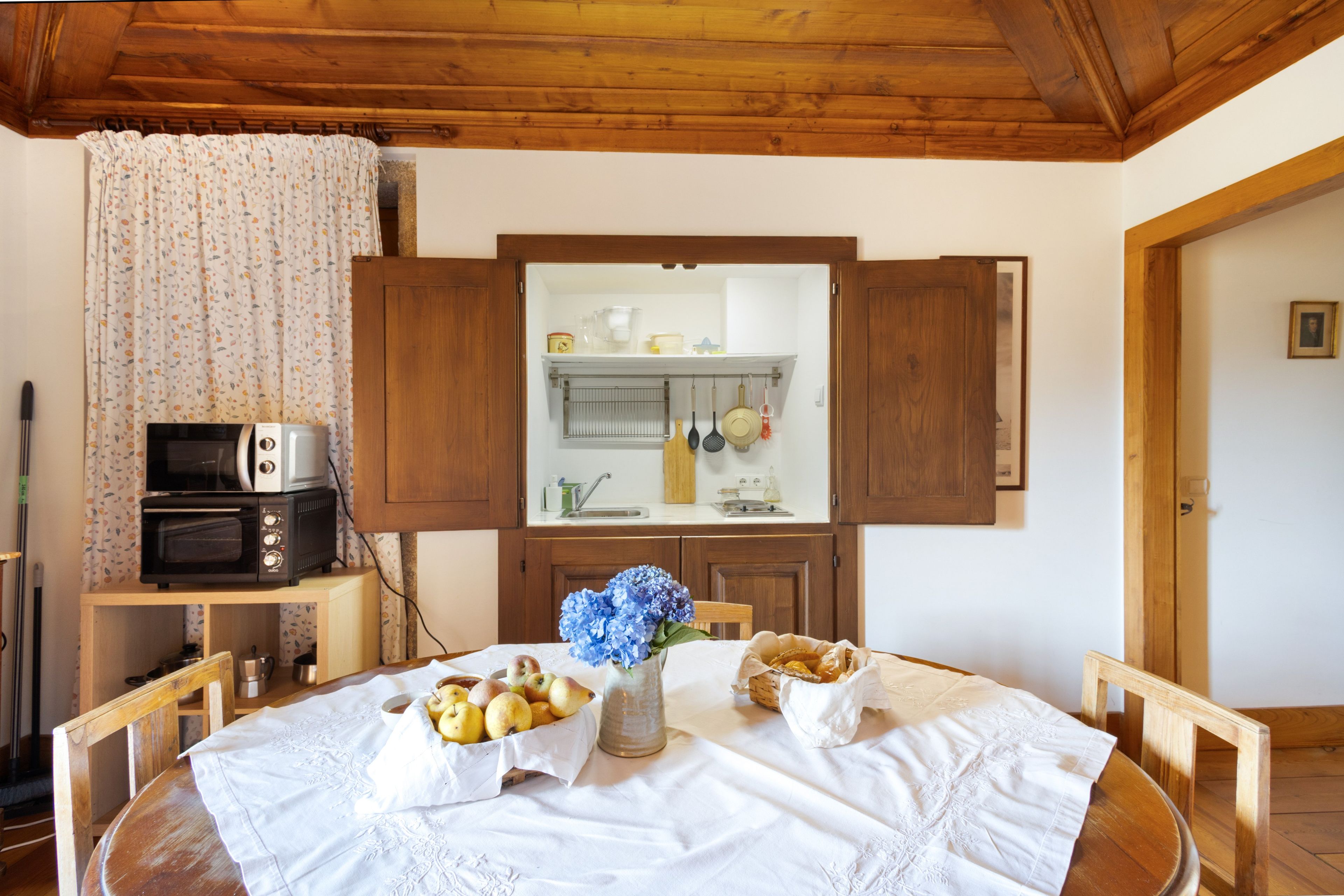 Un comedor con una mesa de madera preparada para dos personas, un florero con flores y una cocina visible a través de un mueble abierto.