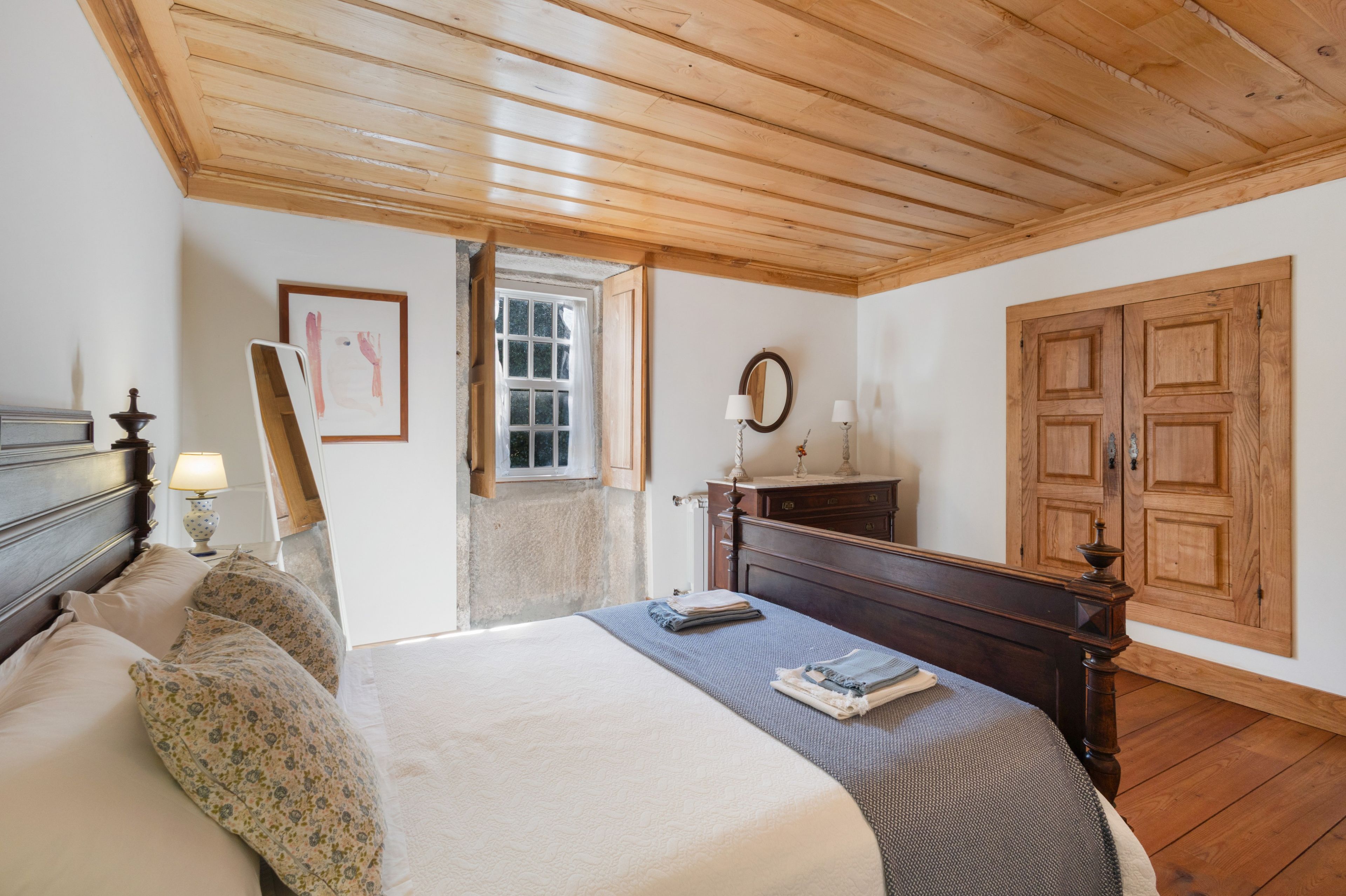 A bedroom with a wooden ceiling, a bed with a headboard, a nightstand with a lamp, a framed picture on the wall, a window, and a wooden wardrobe.